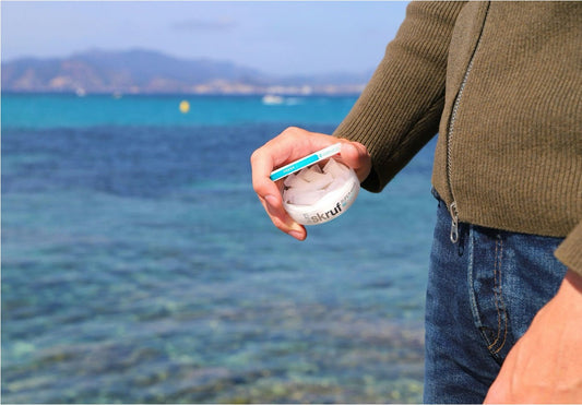 A hand holding an open white can of Skruf Super White Slim nicotine pouches, with a blue sea and coastline in the background