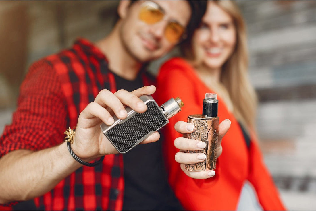 A young couple holding two box-mod vape devices toward the camera