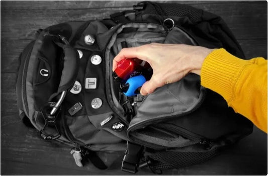 An overhead shot of a person placing two colorful vape devices into the main compartment of a grey backpack