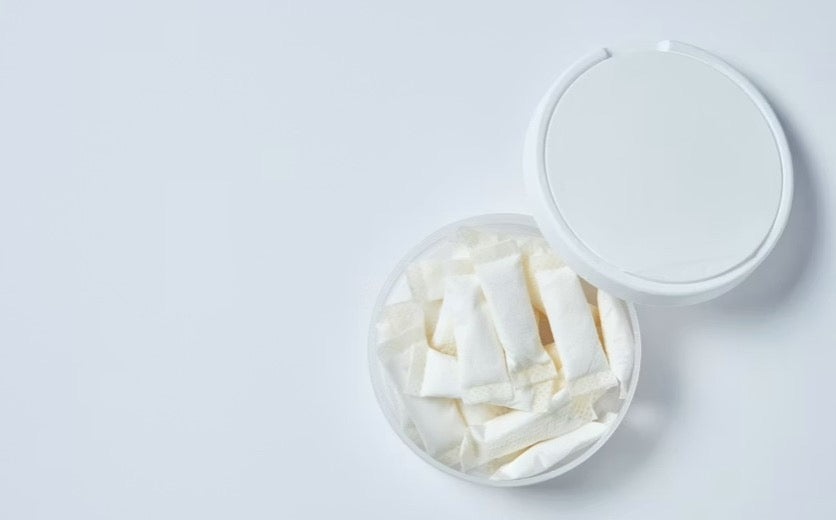 Open white tin filled with white nicotine pouches, lid resting beside it on a light background