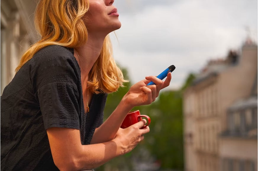 Young woman vaping outside a building, vape device in one hand and a cup of coffee in the other
